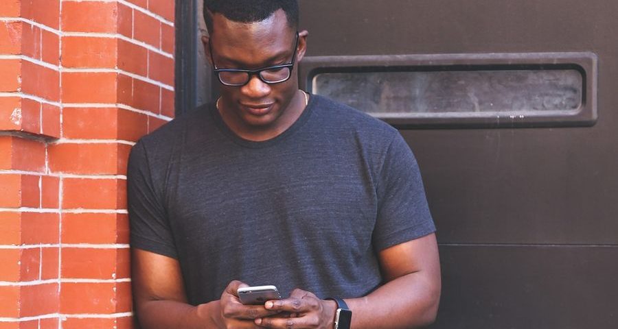 A young black man in glasses and a grey t-shirt looks at his phone, leaning against a red brick wall & brown door