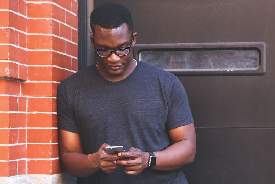 A young black man in glasses and a grey t-shirt looks at his phone, leaning against a red brick wall & brown door