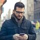 A man with a beard and glasses looks down at his phone on a busy street. By Photo by Andrea Piacquadio on Pexels