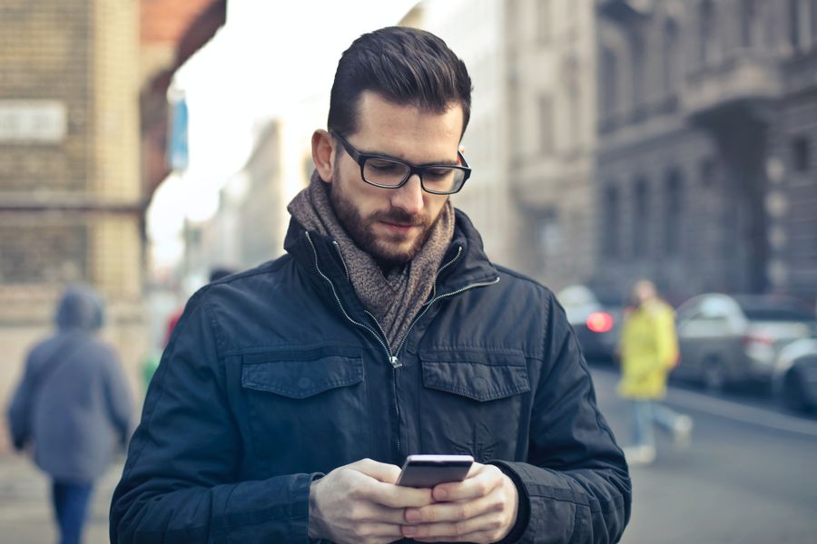 A man with a beard and glasses looks down at his phone on a busy street. By Photo by Andrea Piacquadio on Pexels