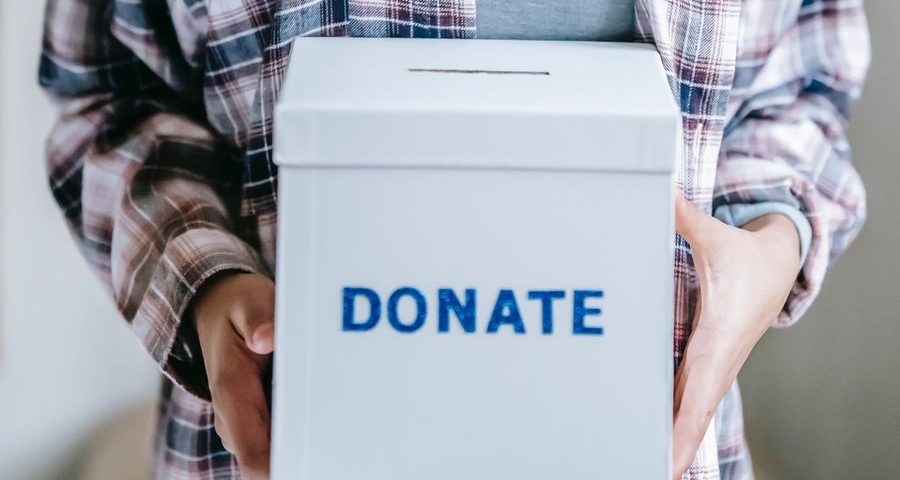 A woman's hands hold a white box with the word Donate on it in blue. By Liza Summer on Pexels