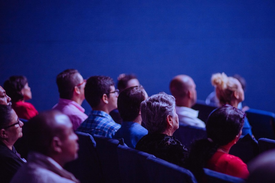 People sit in a darkened audience with a blue background. By Luis Quintero on Pexels