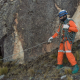A man dressed in safety helmet, goggles and orange protective clothing carries a rope and walks through a rocky landscape, holding onto a rope.
