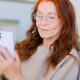 An older woman with long red hair and gold-rimmed glasses sits on a cream-coloured sofa, holding up her phone as she looks at the screen. By Anna Shvets on Pexels