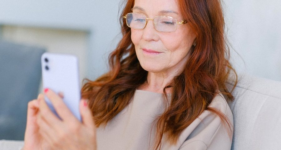 An older woman with long red hair and gold-rimmed glasses sits on a cream-coloured sofa, holding up her phone as she looks at the screen. By Anna Shvets on Pexels
