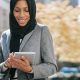 A woman in a black headscarf and grey jacket smiles as she looks at a tablet. By Ono Kosuki on Pexels