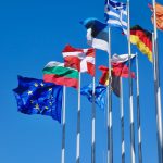 Flags from across the EU plus the EU flag fly against a blue sky. By Antoine Schibler on Unsplash