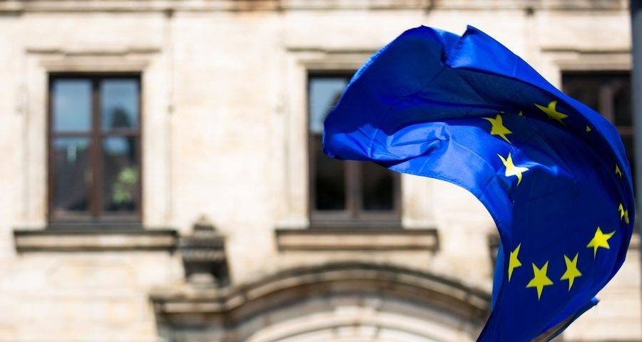 An EU flag flaps in the wind with a stone building in the background. By Markus Spiske on Unsplash