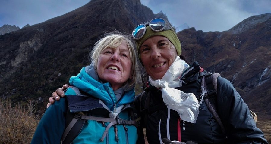 Two older women in walking gear pose for the camera in front of a mountain