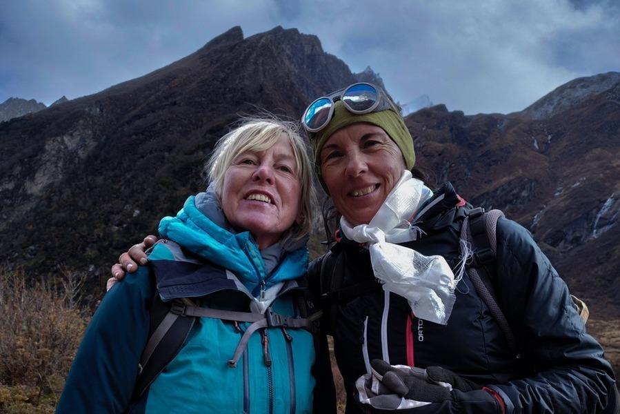 Two older women in walking gear pose for the camera in front of a mountain