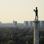 A statue of a standing man holding a bird, with forest and tower blocks in the background, in Pobednik, in Belgrade. By ivalex on Unsplash