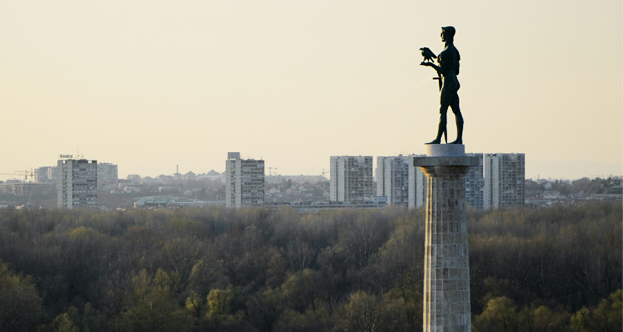 A statue of a standing man holding a bird, with forest and tower blocks in the background, in Pobednik, in Belgrade. By ivalex on Unsplash