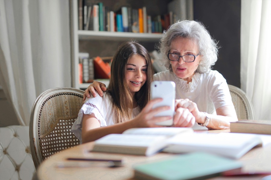 A grandmother sits at a table with her grand daughter who is taking a photo with her phone. By Andrea Piacquadio on pexels