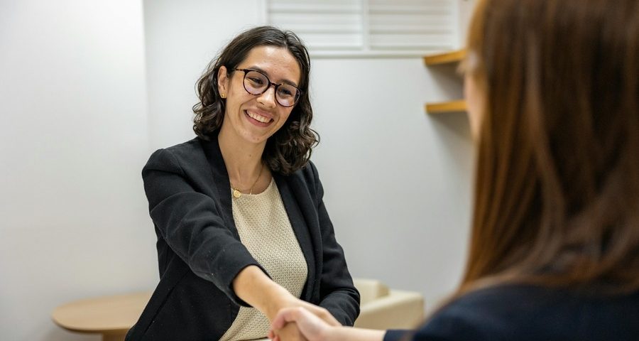 Two women shake hands across a desk in an interview. By Resume Genius on pexels