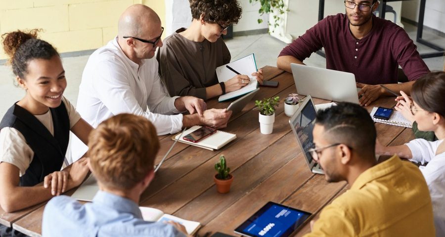 A group of people sit around a table in an office, talking and working. By Fauxels on Pexels.