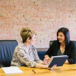 Two women working together talk at a desk. By Amy Hirschi on Unsplash