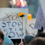 People hold up protest signs and a sunflower in a demonstration again Russia's invasion of Ukraine. By Derek French on Pexels