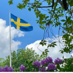 Swedish flag surrounded by green leaves and purple flowers, and a blue sky with big white clouds. By Drahomír Hugo Posteby-Mach via Unsplash