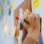 A woman's hand writing on a post-it note on a board. By Getty Images for Unsplash+