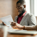 Black man in a suit, sitting at a table in a meeting, looking down at data on sheets of papers_getty Images unsplash+